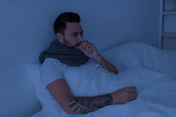 Коронавирус, концепция гриппа. Больной молодой человек кашляет, держит бумажную салфетку у рта, сидит в постели ночью — стоковое фото