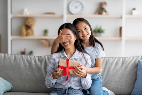 Bonita menina asiática fazendo aniversário surpresa para sua mãe e fechando seus olhos mãe enquanto sentado no sofá — Fotografia de Stock