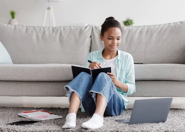 Lachende tiener Afrikaans Amerikaans meisje leerling studies op afstand, schrijft in notebook, zit op de vloer en kijkt naar laptop — Stockfoto