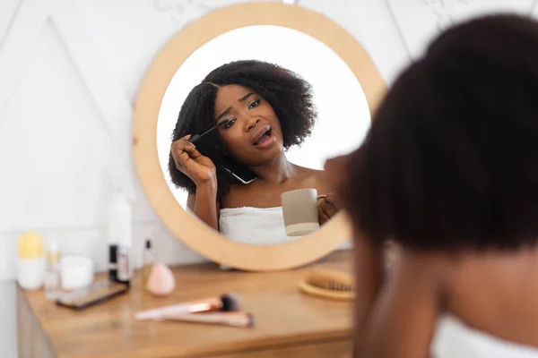 Wanita Afro muda menerapkan maskara pada bulu mata dekat cermin, berbicara di telepon pintar, bergegas untuk bekerja, terlambat di rumah — Stok Foto