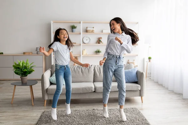 Juguetona madre coreana y bonita hija bailando al son de la música y saltando en casa, disfrutando de su canción favorita juntos — Foto de Stock