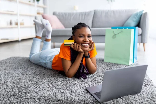 Afro signora con carta di credito per il pagamento a distanza, shopping online via computer portatile, sdraiato sul pavimento con sacchetti regalo a casa — Foto Stock