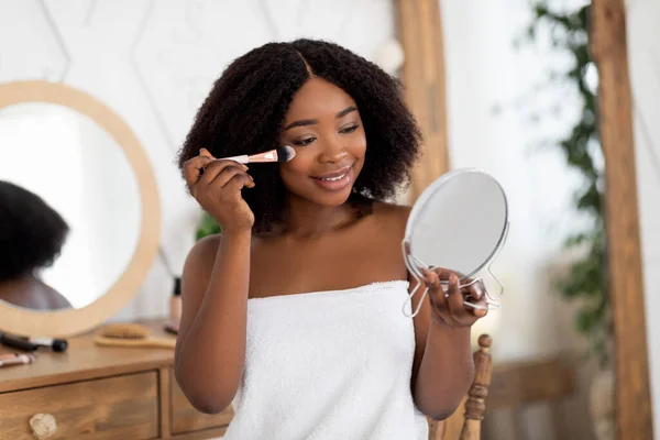 Kosmetické rutiny a make-up. Smyslná mladá Afro žena nanáší prášek nebo červenat, drží zrcadlo doma — Stock fotografie