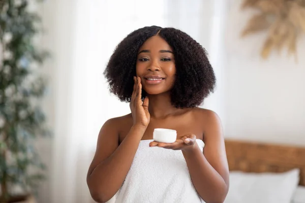 Αισθησιακό νεαρή Afro γυναίκα εφαρμογή κρέμα προσώπου, στέκεται σε πετσέτα μπάνιου, απολαμβάνοντας την οικιακή διαδικασία spa σε εσωτερικούς χώρους — Φωτογραφία Αρχείου