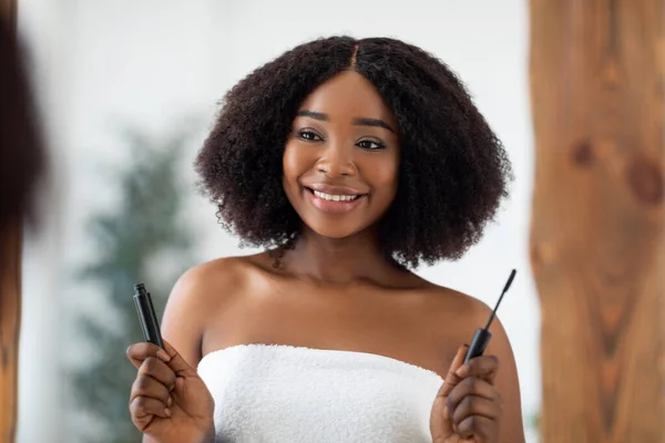 Portrét krásné mladé Afro žena drží řasenku před zrcadlem, pomocí dekorativní make-up doma — Stock fotografie