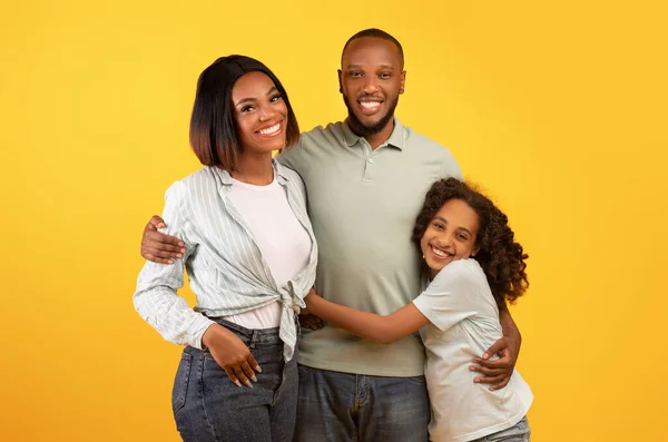 Retrato de família. Felizes pais afro-americanos e linda filha abraçando e posando sobre fundo amarelo — Fotografia de Stock