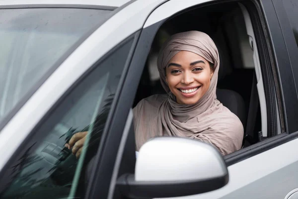 Alegre islámico africano americano señora conductor en hijab se sienta en nuevo coche en el volante y mira a la cámara — Foto de Stock
