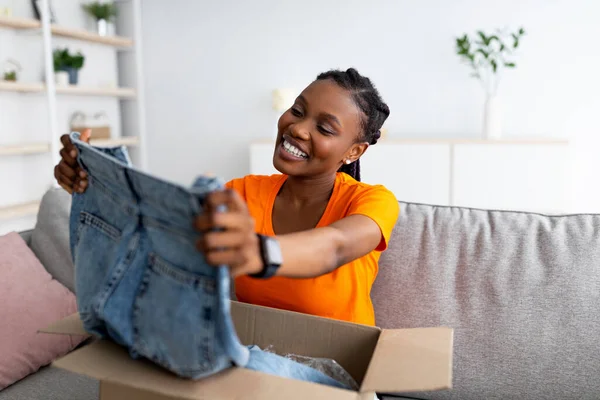Παραγγέλνω ρούχα online. Χαμογελώντας μαύρο θηλυκό πελάτη άνοιγμα δεμάτων κουτί, κοιτάζοντας νέα τζιν στον καναπέ στο σπίτι — Φωτογραφία Αρχείου