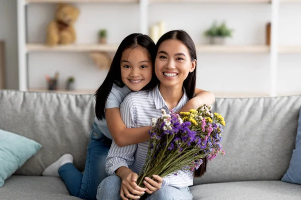 Best wishes on Women Day. Happy asian girl greeting mom and giving her flowers and embracing on sofa