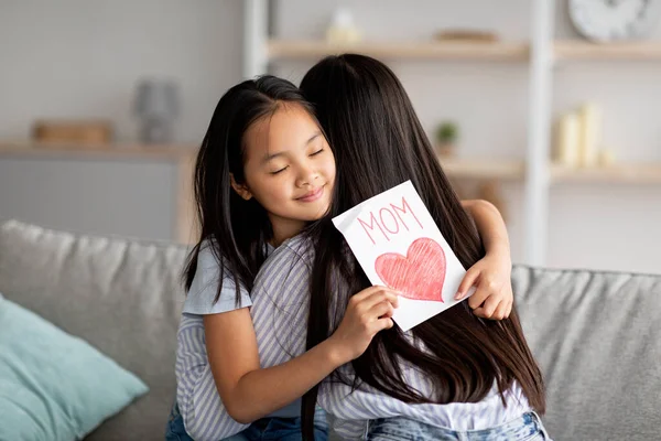 Linda chica asiática abrazando a su mamá, felicitándola con el Día de las Madres, sosteniendo la tarjeta de felicitación, sentada en el sofá — Foto de Stock