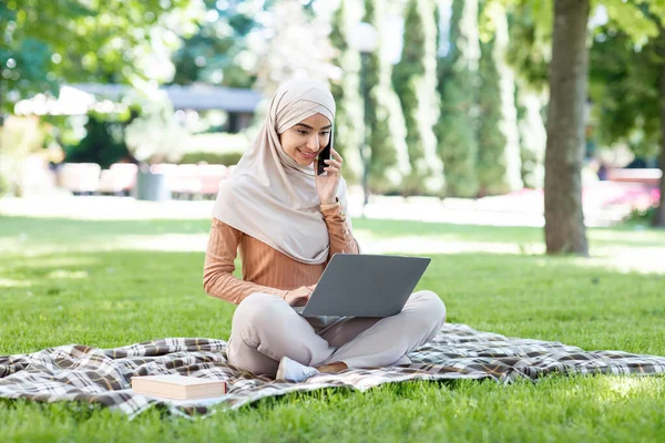 Alegre joven árabe musulmana mujer en hijab utiliza el ordenador portátil y teléfono inteligente para comunicarse con el cliente — Foto de Stock