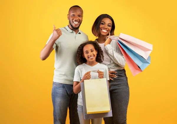 Concept Black Friday. Heureux parents noirs et jolie fille tenant des sacs à provisions, homme montrant pouce vers le haut — Photo