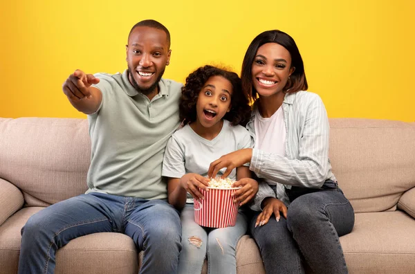 Excited black parents and their little daughter eating popcorn and watching TV, sitting on couch over yellow background