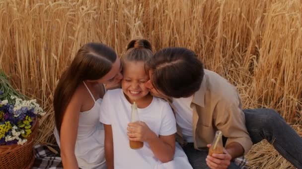 Любящие родители целуют свою дочь во время пикника на пшеничном поле вместе — стоковое видео
