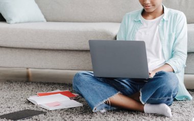 Ciddi bir Afro-Amerikan kızı uzaktan çalışıyor, dizüstü bilgisayarda yazıyor, evinde yerde oturuyor.