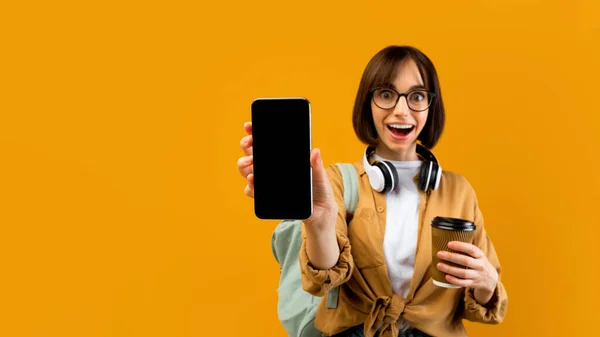 Эмоциональная студентка с пустым экраном на смартфоне, позирующая с рюкзаком и кофе на желтом фоне — стоковое фото