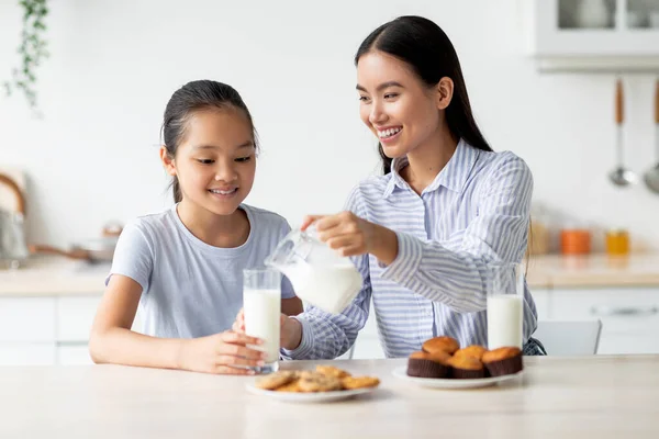 Счастливая азиатская мама и дочь едят закуски и пьют молоко в кухонном интерьере, кусают вместе — стоковое фото