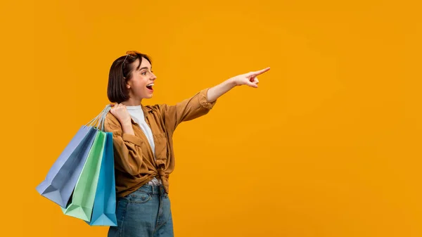 Vaya, míralo. Emocionada dama señalando con el dedo el espacio libre y llevando coloridas bolsas de compras, fondo amarillo — Foto de Stock