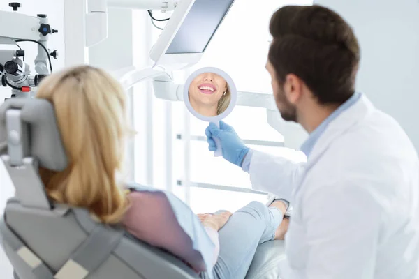 Homem barbudo dentista segurando espelho para paciente sorridente — Fotografia de Stock