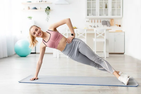 Feliz caucásico hermosa milenaria rubia fitness blogger dama haciendo ejercicio lado tablón en la estera — Foto de Stock