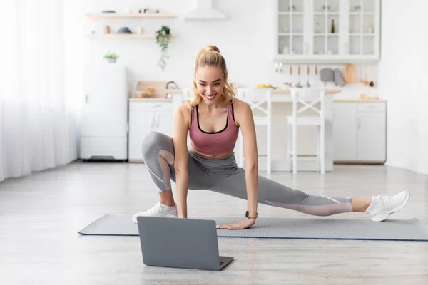 Feliz caucásico hermosa joven rubia atlética haciendo ejercicios de estiramiento de piernas y viendo entrenamiento en línea — Foto de Stock