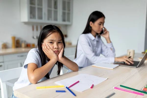 Asiatin arbeitet von zu Hause aus am Laptop, gelangweilte Tochter sitzt neben ihr, lehnt sich an ihre Hände und blickt in die Kamera — Stockfoto