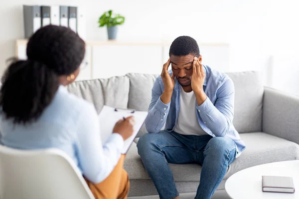 Deprimerad svart man gnuggar sina tempel, konsulterar psykolog, sitter på soffan på rådgivningsbyrån — Stockfoto