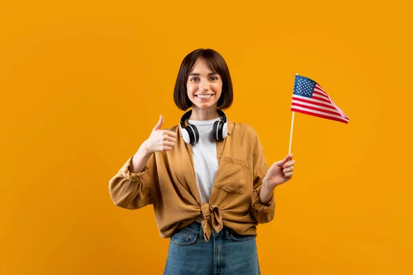 Happy lady tonen duim omhoog en Amerikaanse vlag, poseren met koptelefoon op de hals over gele achtergrond, kopieer ruimte — Stockfoto