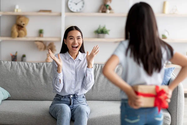 Liten flicka gömmer inslagna presentförpackning för semester bakom ryggen, gratulera sin förvånade mamma med födelsedag hemma — Stockfoto