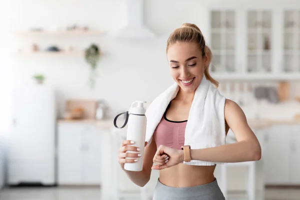 Lächelnde kaukasische hübsche junge blonde Frau überprüft Herzschlag auf Fitness-Tracker und trinkt Wasser nach dem Training — Stockfoto