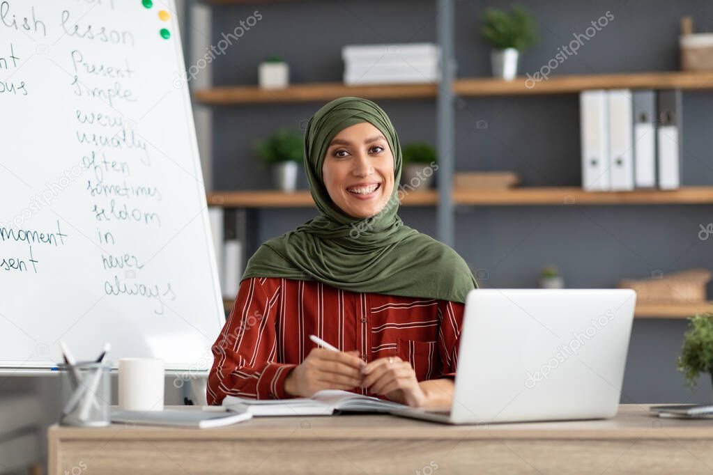 Islamic teacher in headscarf sitting at desk looking at camera