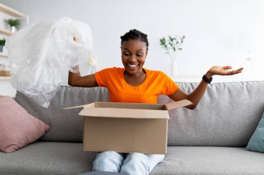 Heyecanlı siyahi kadın paket alıyor, karton paketleri açıyor, plastik ambalaj kağıdı tutuyor, evdeki kanepede oturuyor.