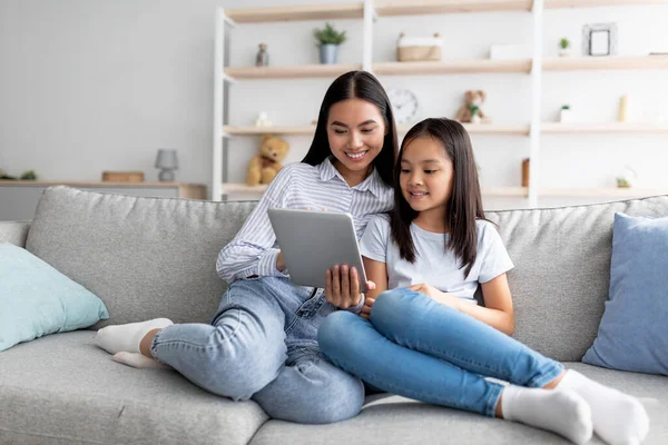 Ung asiatisk mamma och dotter som sitter på soffan och använder digitala surfplattor, tittar på videor eller surfar internet — Stockfoto