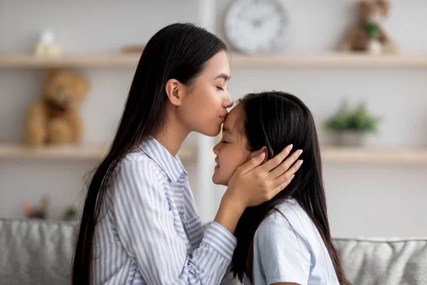 Las madres aman. cariñosa asiática mamá besos su hija en la frente mientras pasando tiempo juntos en casa, lado ver —  Fotos de Stock