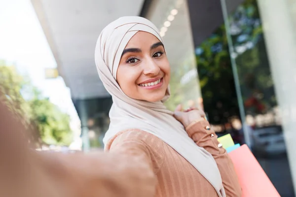 Veselý rád pěkný tisíciletý arabský kupující blogger v hidžábu se spoustou tašek dělá selfie na zařízení — Stock fotografie