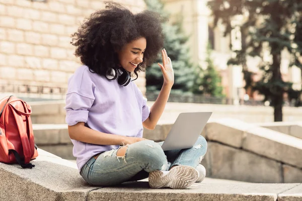 Negro estudiante chica vídeo llamadas usando portátil ondeando mano al aire libre — Foto de Stock