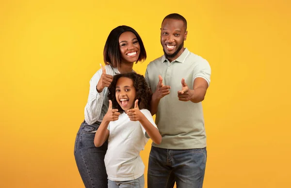 Hé, jij daar. Opgewonden zwarte ouders en hun dochter wijzen met de vingers naar de camera, staande over gele achtergrond — Stockfoto