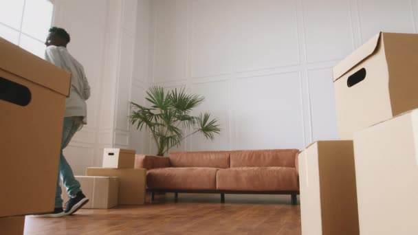 Joven hombre afroamericano positivo bailando con caja en su nuevo apartamento, disfrutando mudarse a casa, sentado en el sofá — Vídeos de Stock