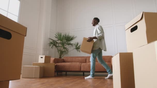 Young happy african american guy moving to new home, carrying box with stuff, resting on sofa, enjoying new apartment — Stock Video