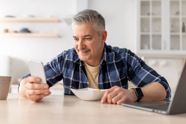 Feliz hombre maduro utilizando el teléfono inteligente mientras desayuna en la mesa de la cocina en casa, espacio para copiar —  Fotos de Stock