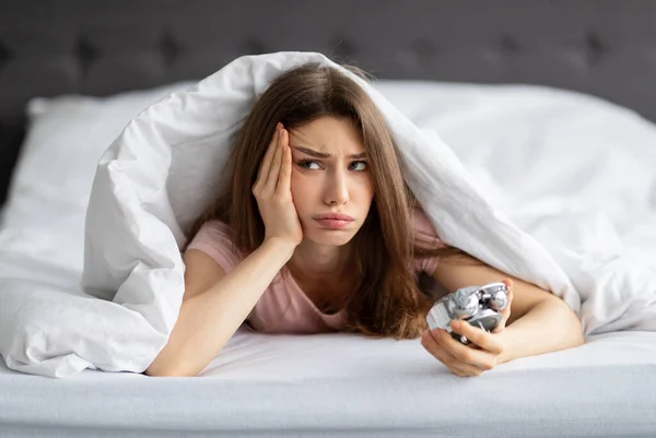 Jovem mulher caucasiana infeliz deitada na cama com despertador, não disposta a levantar-se de manhã — Fotografia de Stock