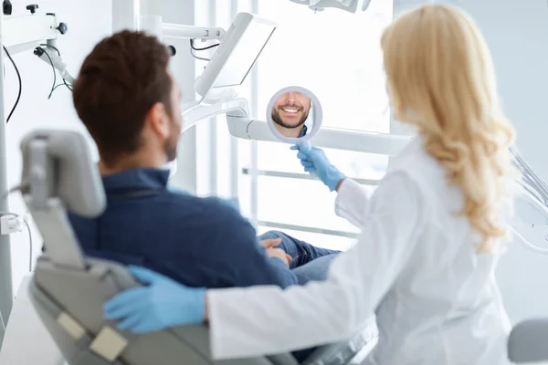웃는 남자 환자를 위해 거울을 들고 있는 암컷 치과 의사 — 스톡 사진
