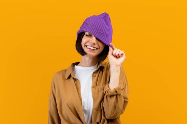 Mor şapkayı indiren, bir gözünü kapatan ve turuncu arka planda gülümseyen mutlu genç kadın.