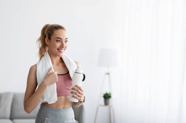 Cheerful european young blonde fitness blogger athlete with bottle of water resting after workout at home clipart