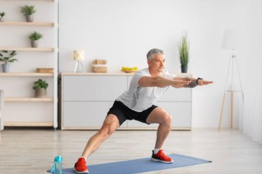 Ev antrenmanı. Aktif, olgun bir adam yan hamleler yapıyor, oturma odasında yoga minderinde bacaklarını açıyor, uzayı kopyalıyor.