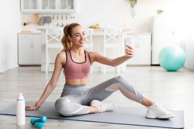 Dostça gülümseyen Kafkasyalı genç sarışın fitness blogcusu telefon webcam 'ine bak ve canlı yayında egzersiz yap.