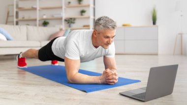 Mutlu yetişkin adam egzersiz yapıyor ve dizüstü bilgisayarda video izliyor oturma odasında çalışırken kalas yapıyor.