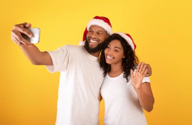 Birlikte anılar yaratıyoruz. Noel Baba şapkalı Afro-Amerikan çift sarı arka planda selfie çekiyorlar.