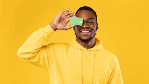Czarny facet zakrywający oko kartą kredytową, żółte tło — Zdjęcie stockowe