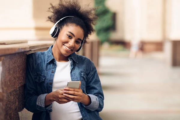 Nero studente ragazza holding smartphone indossare wireless auricolari in piedi all'aperto — Foto Stock